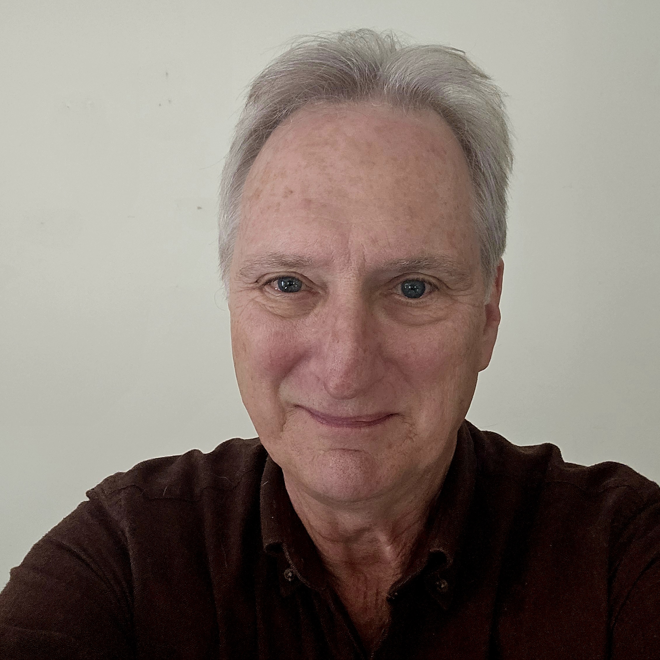 Portrait of a gray-haired man in a dark shirt smiling gently at the camera in a close-up selfie against a plain light background.